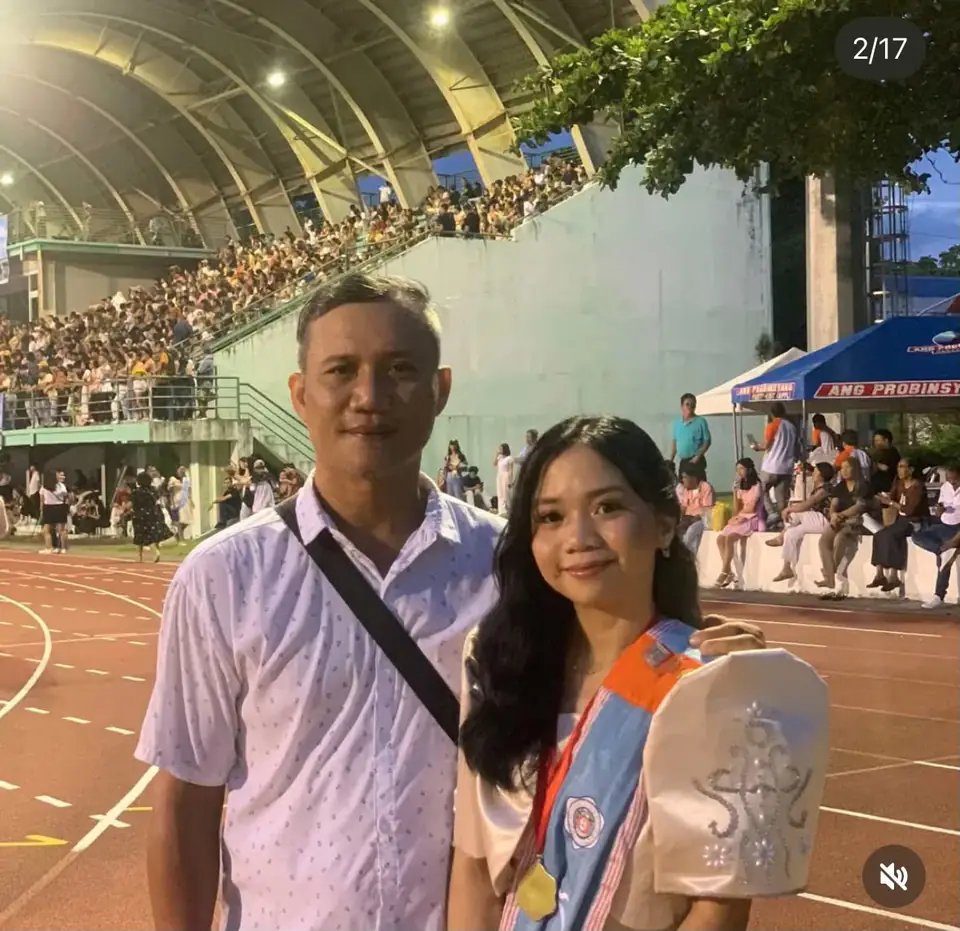 Daniele in her graduation sash and medal, standing beside a man on a stadium running track