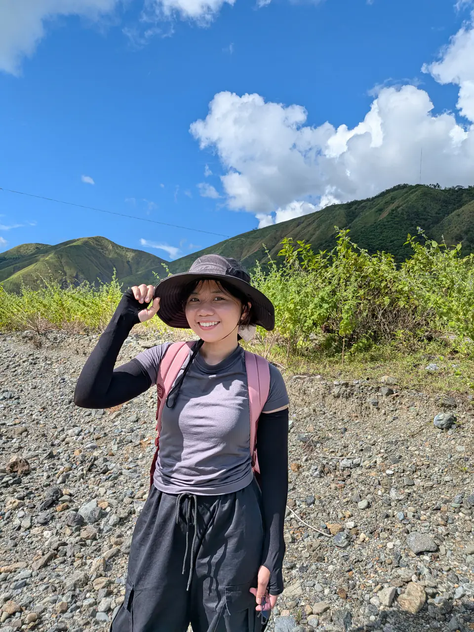 Daniele in a sun hat and backpack on a rocky trail, with green mountains and blue sky behind her